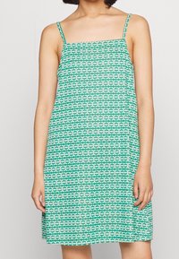 Zelené bezrukoé šaty s geometrickým bílým vzorem, vyrobené z lehkého materiálu, s tenkými ramínky a volným střihem.