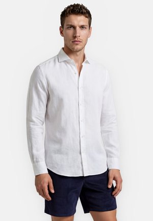 Mand iført hvid skjorte med lange ærmer og mørkeblå shorts, stående mod en ensfarvet lys baggrund.