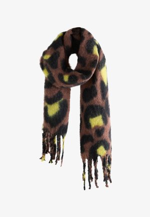Person trägt eine beige Jacke und einen flauschigen, mehrfarbigen Schal mit schwarzen, braunen und gelben Mustern, sitzt mit den Händen auf dunkler Hose.