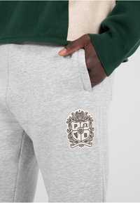 Šedé tepláky s vyšitým emblémem, který zobrazuje design štítu. Měkká tkanina s hladkou strukturou, navržená pro pohodlí.