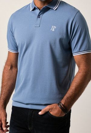 Mann trägt ein blaues Poloshirt mit weiß gestreiftem Kragen und Ärmeln, schwarze Hose und ein schwarzes Armband, Hand in der Tasche.