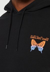 Felpa nera con una grafica di una farfalla in arancione e blu. Il testo sopra recita "la luce di una luna piena" e "magia della luce della luna". Cappuccio regolabile con cordino.