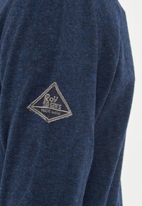Maglione blu navy realizzato in un tessuto a maglia testurizzato. Presenta una toppa grigia triangolare con la scritta "Roy" e "Trade Mark" sulla manica sinistra.