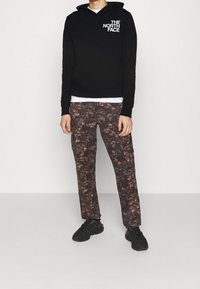 Svart hoodie med vit logga, matchad med mönstrade bruna byxor med lövdesign. Bärs med svarta sportskor. Enkel bakgrund.