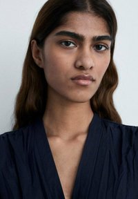 Krupni plan osobe s dugom, valovitom smeškom. Ima istaknute tamne obrve, glatku kožu i nosi tamnoplavu majicu od teksturiranog materijala.