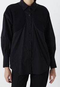 Persona che indossa una camicia nera con bottoni e toppe circolari testurizzate sul petto superiore e maniche lunghe, abbinata a pantaloni neri.
