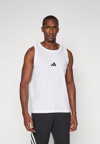 Bela majica brez rokavov iz bombaža z okroglo izrezom in črnim logotipom Adidas na prsih, v kombinaciji s temno sivo hlačami z tremi črtami.
