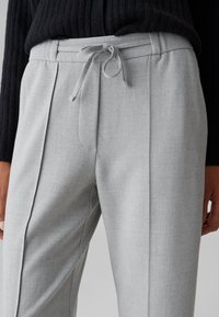 Helle graue maßgeschneiderte Hose mit glatter Textur, ausgestattet mit einem Kordelzugbund und vorderen Falten für zusätzliche Details.