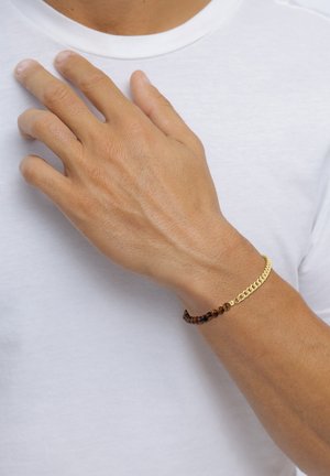 Hand mit einem gold- und braunperligen Armband, die auf der weißen Hemdbrust ruht.