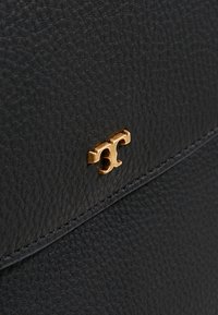 Surface en cuir texturé noir avec un petit logo double-T doré fixé près d’un bord cousu.