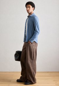 Camisa azul de mangas compridas com botões, calças castanhas de perna larga e sapatos pretos. Modelo segura uma mala preta texturada. Fundo minimalista.