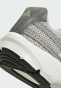 Gros plan sur le talon d'une basket Adidas gris et blanc avec une tige en maille, des superpositions synthétiques et une semelle en caoutchouc noire texturée.