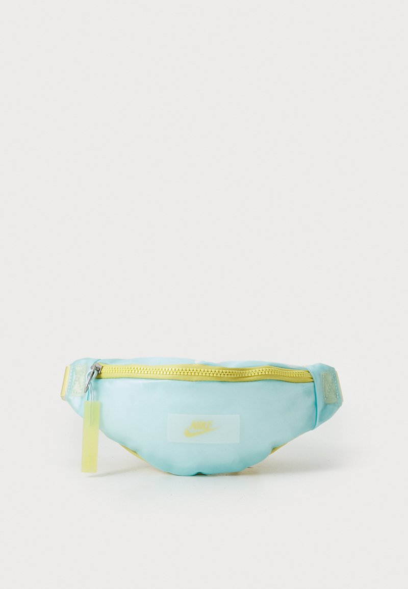 Gaiši zils fanny pack ar spīdīgu apdari, dzeltenu rāvējslēdzēju un baltu Nike logotipa uzšuvumu. Tajā ir pievienota dzeltena birka pie rāvējslēdzēja.