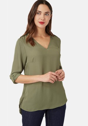 Mujer vestida con blusa verde oliva de escote en V y mangas arremangadas, y vaqueros oscuros, de pie contra un fondo claro y liso.
