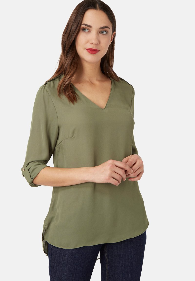 Mujer vestida con blusa verde oliva de escote en V y mangas arremangadas, y vaqueros oscuros, de pie contra un fondo claro y liso.