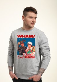 Uomo che indossa una felpa grigia con la grafica di Wham! Last Christmas, raffigurante due uomini in costumi di Natale che tengono scatole regalo.