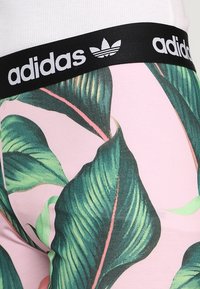 Legíny Adidas s černým páskem s logem; růžová látka má velké zelené listové vzory; hladká textura a těsný střih.