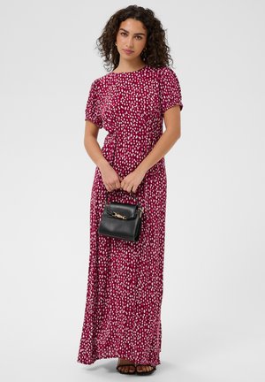 Kobieta o kręconych ciemnych włosach, ubrana w długą czerwoną sukienkę w białe kropki, trzymająca małą czarną torebkę ze złotymi detalami.