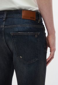 Sötét farmernadrág bőr BOSS logóval ellátott címkével. Jellemzői a klasszikus ötlovas zsebes kialakítás és a látható varrási részletek.