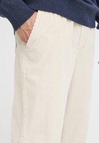 Beżowe spodnie sztruksowe z pionowymi prążkami, z przednimi kieszeniami i elastycznym pasem, częściowo widoczne obok ciemnoniebieskiego swetra.