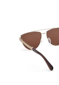 Gafas de sol aviador con montura dorada y lentes marrones, que cuentan con un puente superior de metal, almohadillas nasales transparentes y patillas de plástico marrón oscuro.