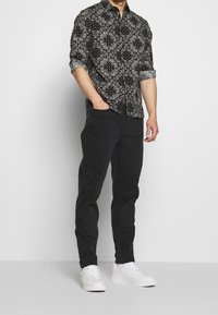 Černá košile s uzorhem a paisley designem, spárovaná s černými kalhotami a bílými teniskami. Košile má ohrnuté rukávy a voln ý střih.