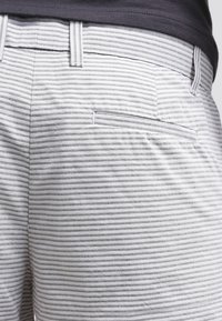 Gros plan sur une poche de short rayé gris et blanc avec passants de ceinture portée par une personne en chemise sombre.