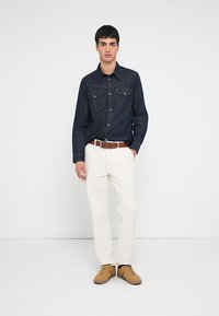 Ciemnoniebieska koszula jeansowa z dwiema przednimi kieszeniami, jasne spodnie, brązowy pasek i beżowe trampki. Prosty i czysty design.