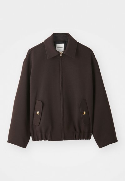 Donkerbruine jas met een rits aan de voorkant, wijde kraag, twee opgestikte zakken met gouden knopen en een elastische onderrand. Gemaakt van een gladde stof.