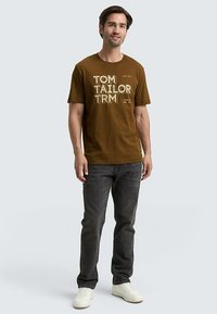 Brązowy bawełniany T-shirt z białym i beżowym napisem "TOM TAILOR TRM." Model stoi na tle jasnego tła, ubrany w ciemne jeansy i białe sneakersy.