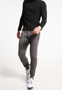 Pantalones deportivos grises hechos de tela elástica con piernas cónicas, cremalleras laterales, combinados con una camiseta henley negra de manga larga y zapatillas blancas.