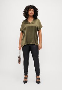 Blusa di raso verde oliva con scollo a V e maniche corte abbinata a leggings neri attillati e scarpe con tacco alto nere a tracolla.