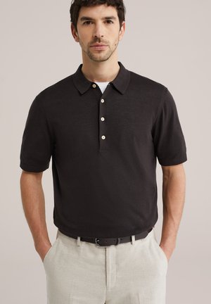 Moški, oblečen v črno polo majico s kratkimi rokavi, bež hlače in črni pas, stoji z rokami v žepih na enobarvnem ozadju.