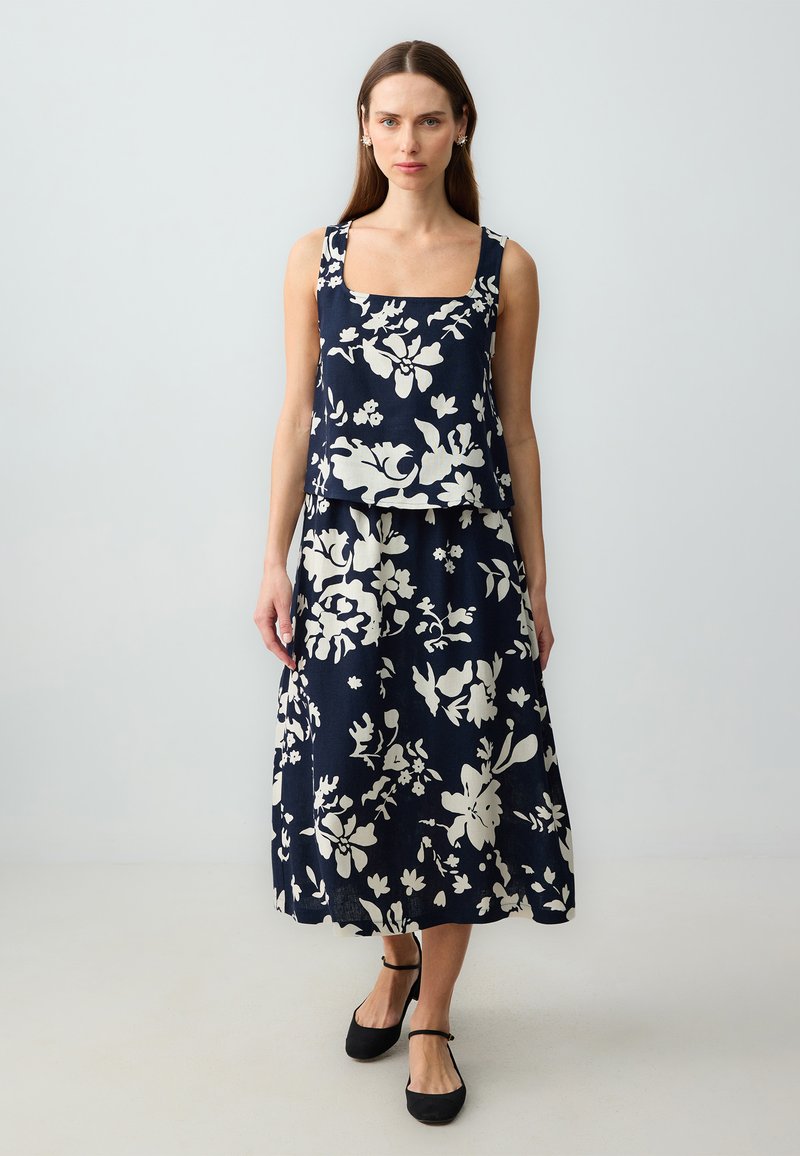 Blumenmuster Kleid im zweiteiligen Design in Marineblau und Weiß. Das Oberteil hat einen quadratischen Ausschnitt, während der Rock unter das Knie fließt. Kombiniert mit schwarzen Schuhen.