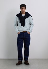 Hombre vistiendo un sudadera gris claro con un suéter azul marino sobre los hombros, jeans azul oscuro, cinturón marrón y botas marrón oscuro, de pie en el interior.