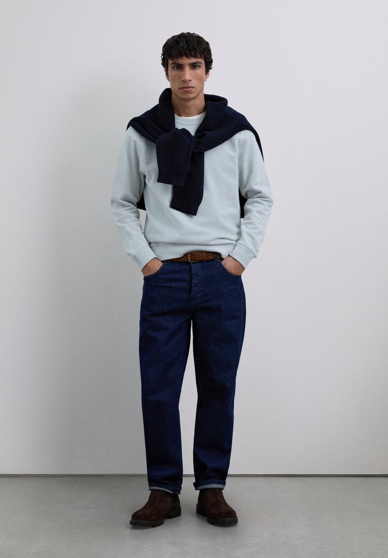 Hombre vistiendo un sudadera gris claro con un suéter azul marino sobre los hombros, jeans azul oscuro, cinturón marrón y botas marrón oscuro, de pie en el interior.