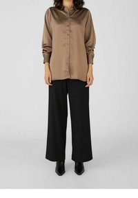 Persona che indossa una camicia a maniche lunghe in satin marrone con bottoni, pantaloni neri a gamba larga e stivali neri con punta a punta, in piedi su un pavimento grigio.