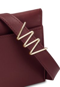 Bordeaux kožená kabelka s širokým popruhem, která má zlatý kovový akcent ve tvaru cik-cak. Hladký povrch s viditelnými detaily šití.