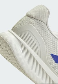 Zapatilla deportiva gris claro con parte superior de malla transpirable, suela de goma texturizada y un acento azul. Presenta un collar acolchado y un diseño aerodinámico.