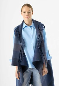 Sciarpa blu navy con bordi frangiati, indossata sopra una camicia blu chiaro con bottoni. La sciarpa presenta leggere fantasie a quadretti e una consistenza morbida.
