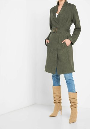 Vrouw draagt een olijfgroene trenchcoat met ceintuur, lichtblauwe jeans en beige slordige hoge laarzen, staand tegen een witte hoekmuur.