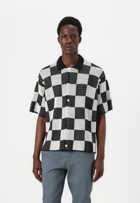 Chemise à manches courtes avec un motif à carreaux noir et blanc, col noir contrastant et fermeture à boutons. Le tissu présente un tissage texturé.