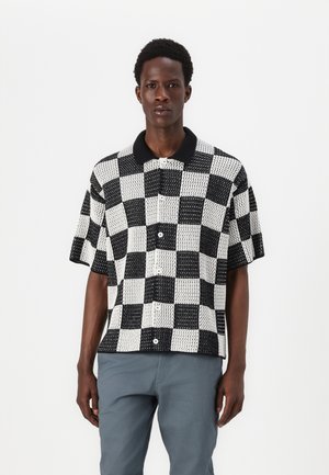 Chemise à manches courtes avec un motif à carreaux noir et blanc, col noir contrastant et fermeture à boutons. Le tissu présente un tissage texturé.