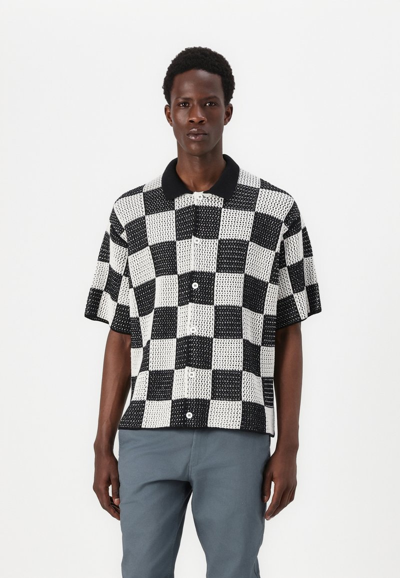 Chemise à manches courtes avec un motif à carreaux noir et blanc, col noir contrastant et fermeture à boutons. Le tissu présente un tissage texturé.