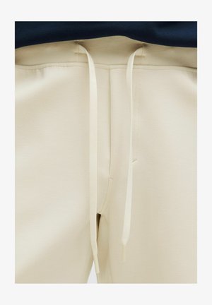 Bézs színű melegítőnadrág, amely elasztikus derékrésszel, fehér húzózsinórokkal, sima textúrával és karcsúsított szabással rendelkezik.