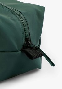 Bolsa verde con cremallera de textura suave, con un tirador y una correa de cremallera negra resistente. El diseño es rectangular con bordes redondeados.