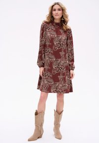 Brązowa sukienka w kwiatowy wzór z długimi rękawami, wysoką szyją i zwiewnym materiałem. Połączona z jasnobeżowymi kowbojkami.