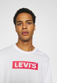 T-shirt bianco in cotone con scollo a girocollo, presenta un logo rettangolare rosso con la scritta "LEVI'S" in caratteri bianchi in grassetto. Texture liscia, design casual.