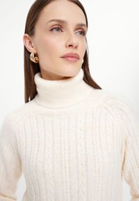 Zachte beige coltrui met een textuur kabelbreipatroon, voorzien van een geribde kraag en manchetten. Model draagt gouden oorringen.