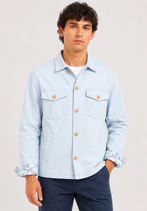 Homme aux cheveux bouclés porte une veste bleu clair boutonnée avec des détails floraux aux poignets et un pantalon bleu foncé, debout devant un fond uni.